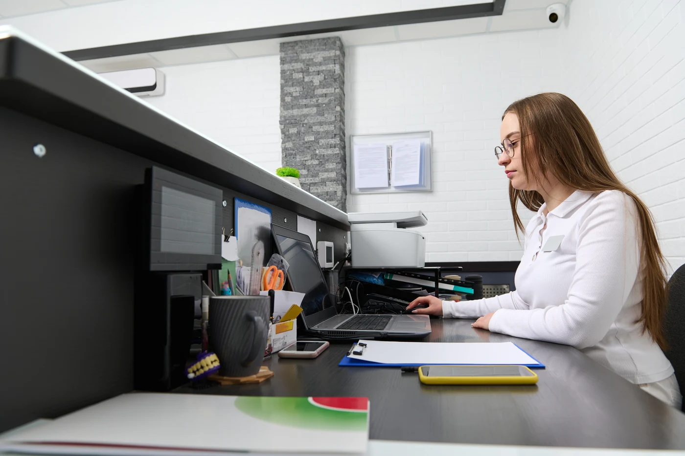 Receptionist working at a laptop beside a printer and office paperwork