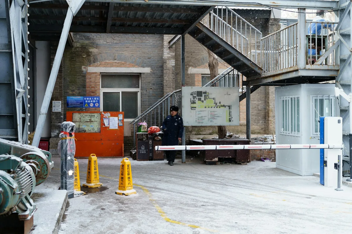 A guard beside a barrier gate and booth at a snow-covered industrial entrance.