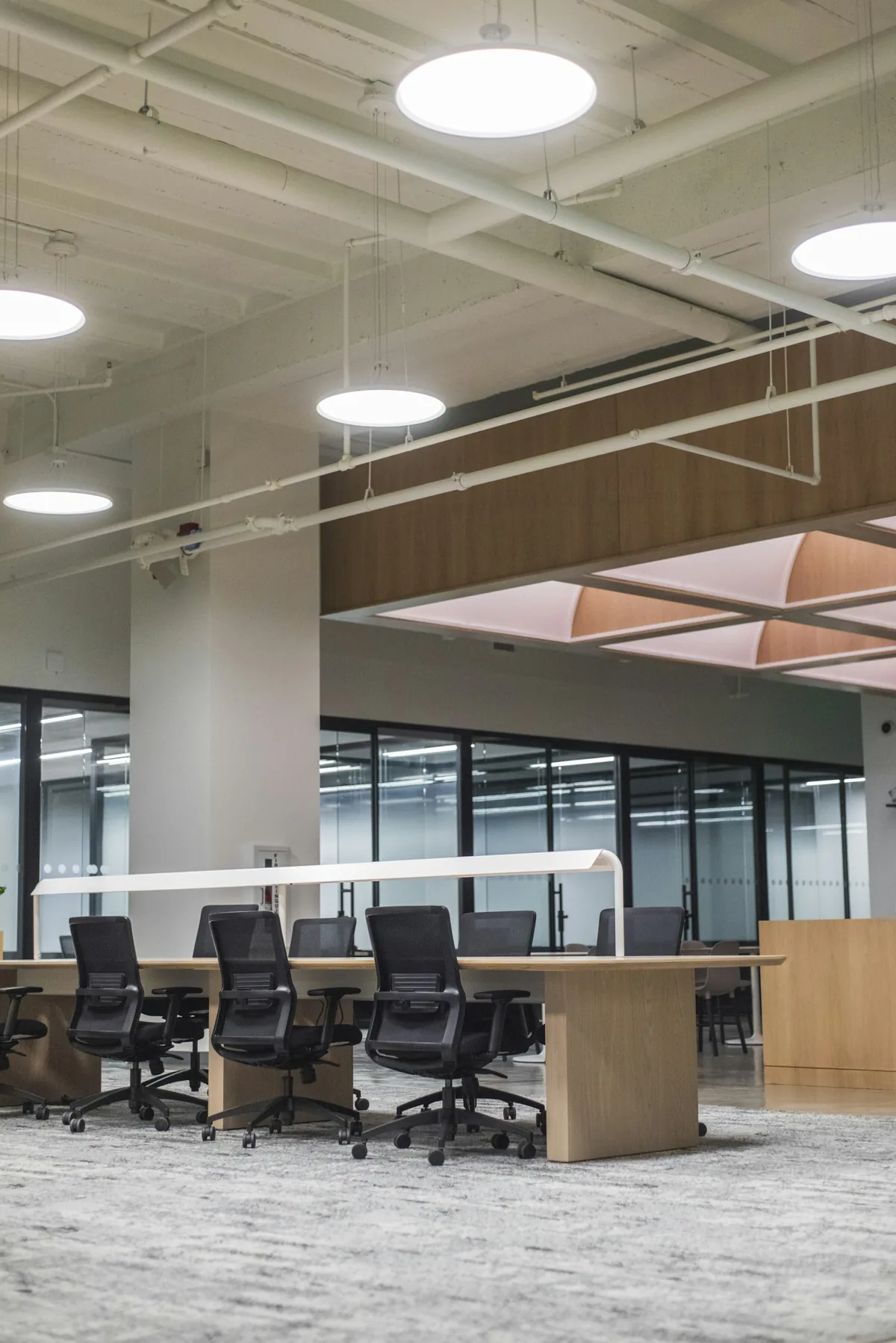 An open office with desks, rolling chairs, glass partitions, and exposed ceiling services.