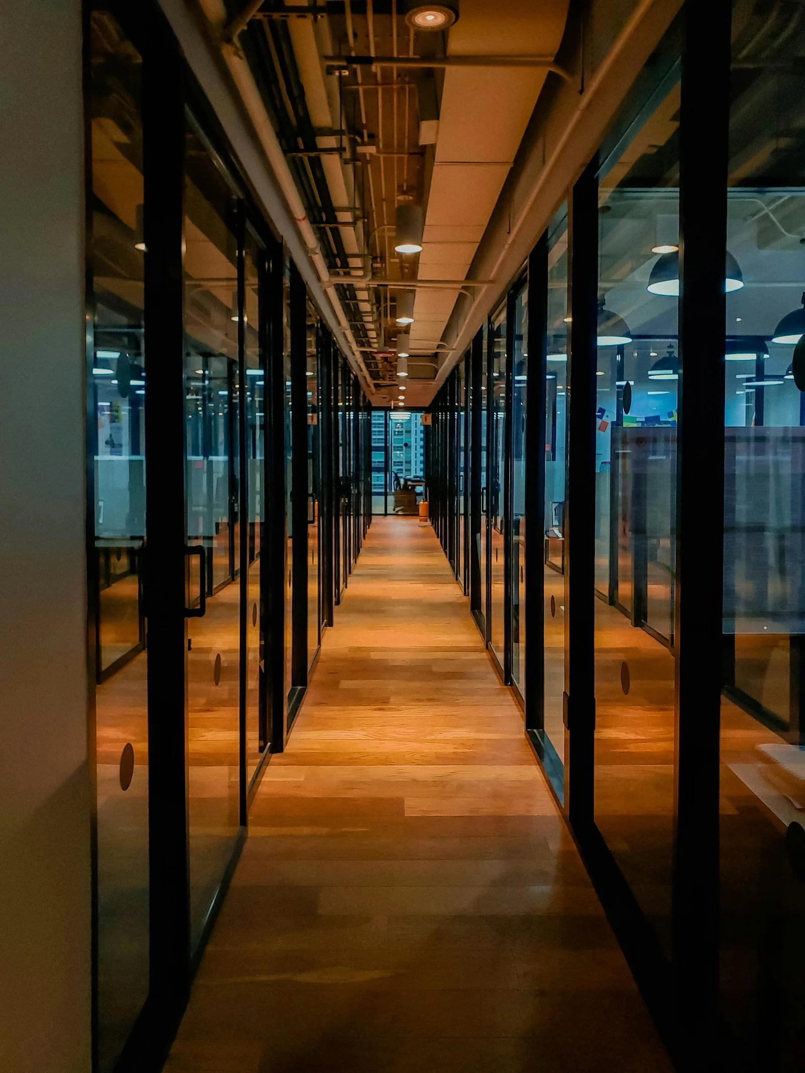 A long office corridor with glass doors, wood flooring, and exposed overhead services.
