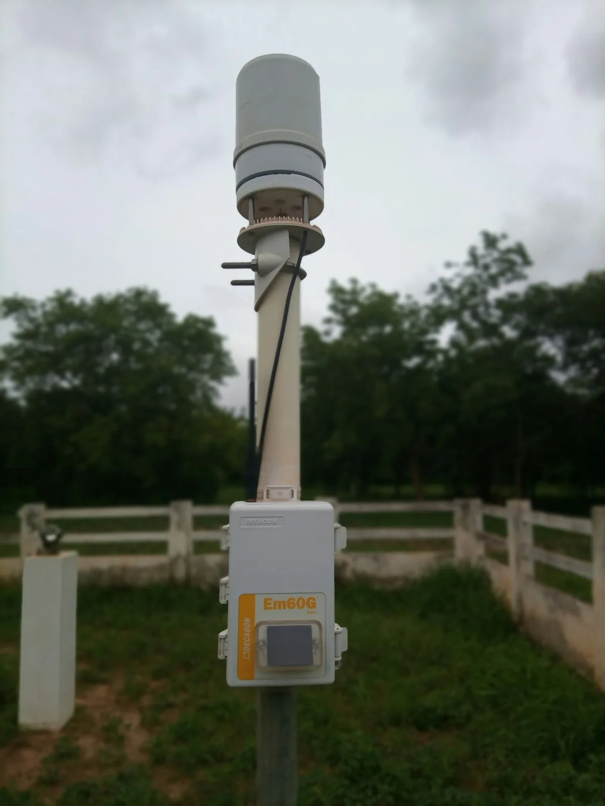 Outdoor environmental instrument mounted on a post beside a fenced green area.