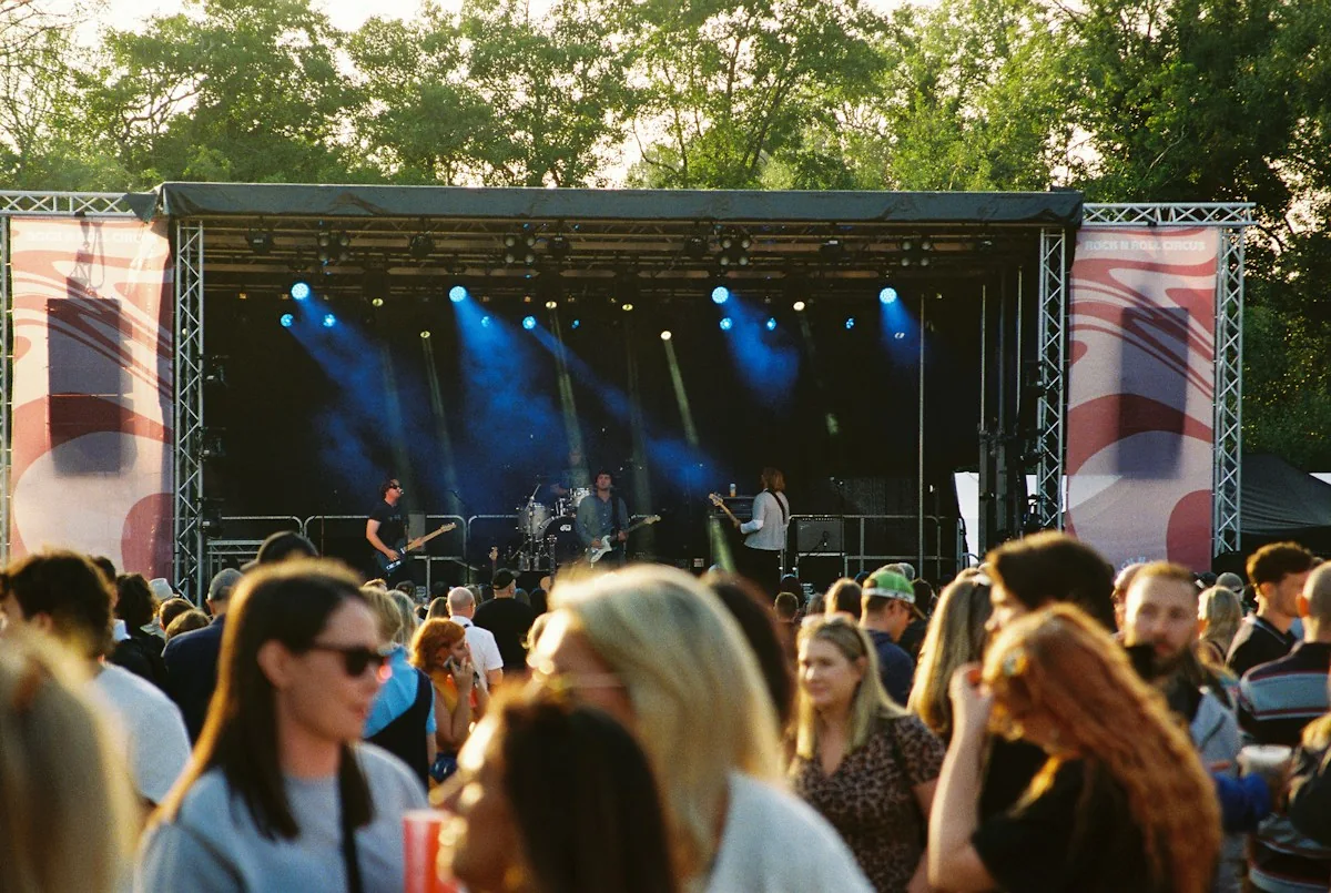 Audience members gather before an outdoor concert stage beneath trees.