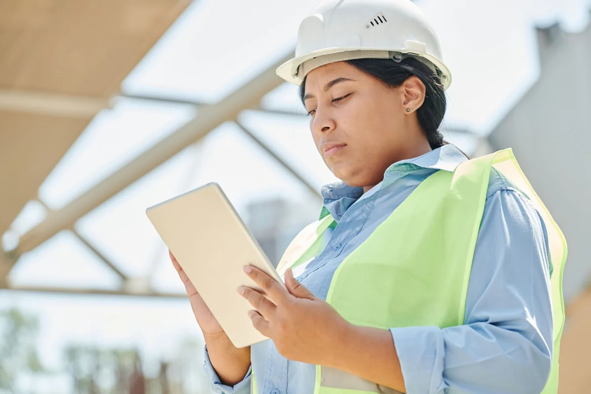 Worker in a white hardhat and high-visibility vest using a tablet beside a building frame.