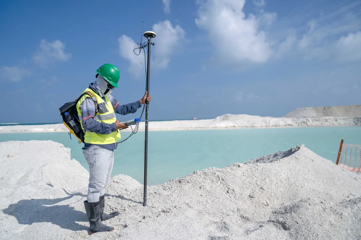 Survey worker holding a positioning receiver on a pole beside a pale excavated shoreline.