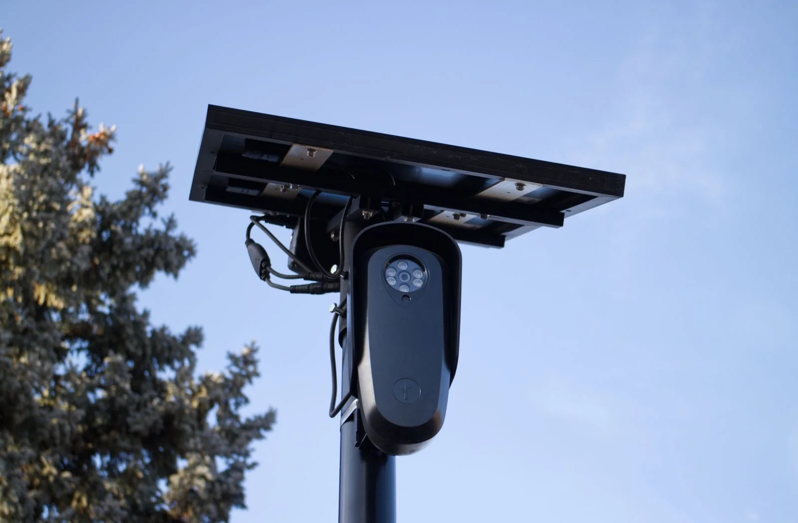 Outdoor surveillance camera beneath a small solar panel on a pole against a blue sky.