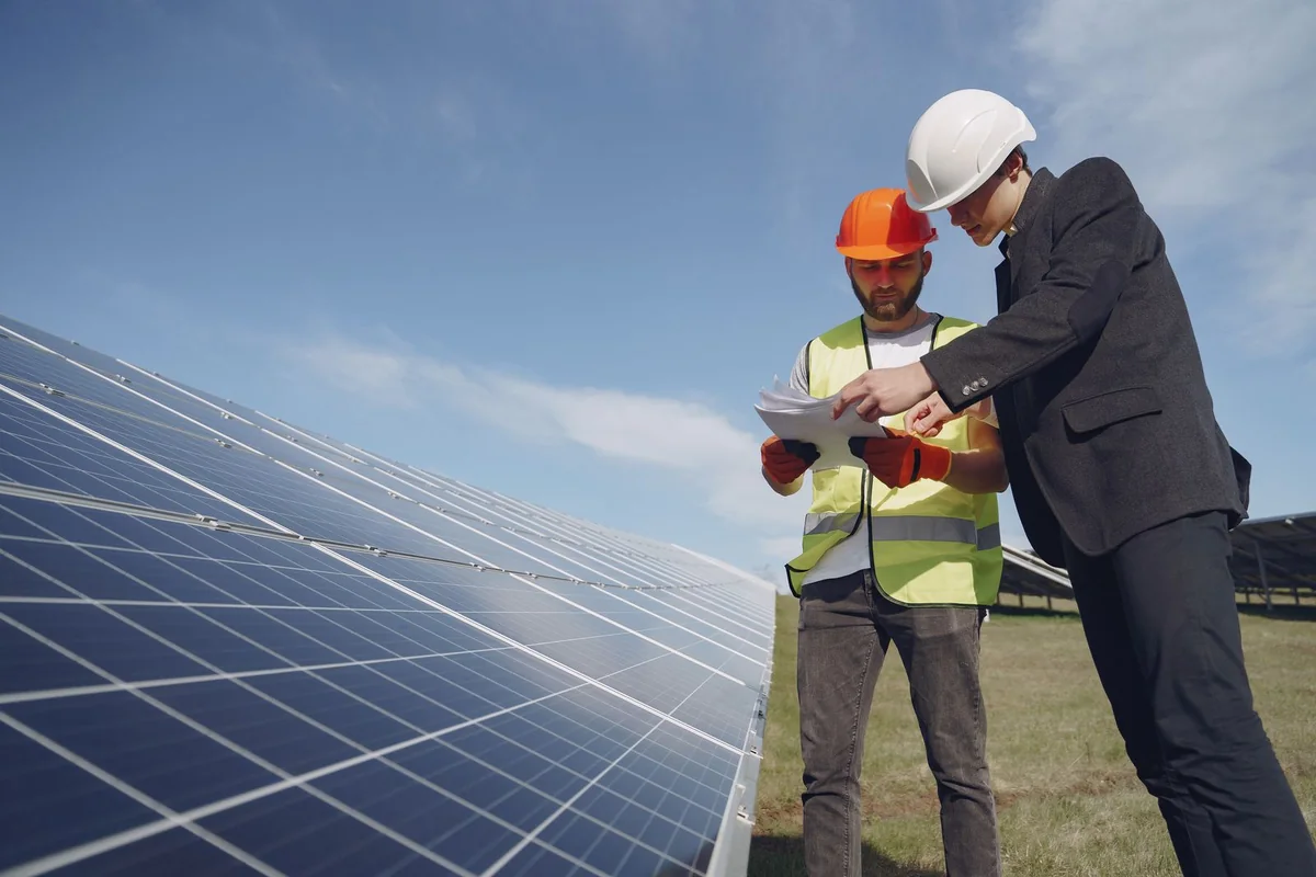 Two people wearing hard hats review paperwork beside an outdoor solar-panel array.