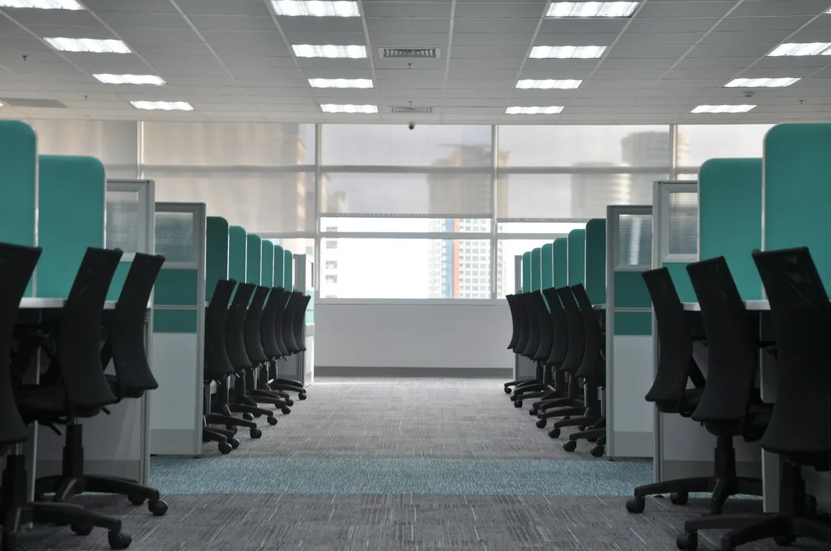 Rows of office cubicles beneath illuminated rectangular ceiling lights.
