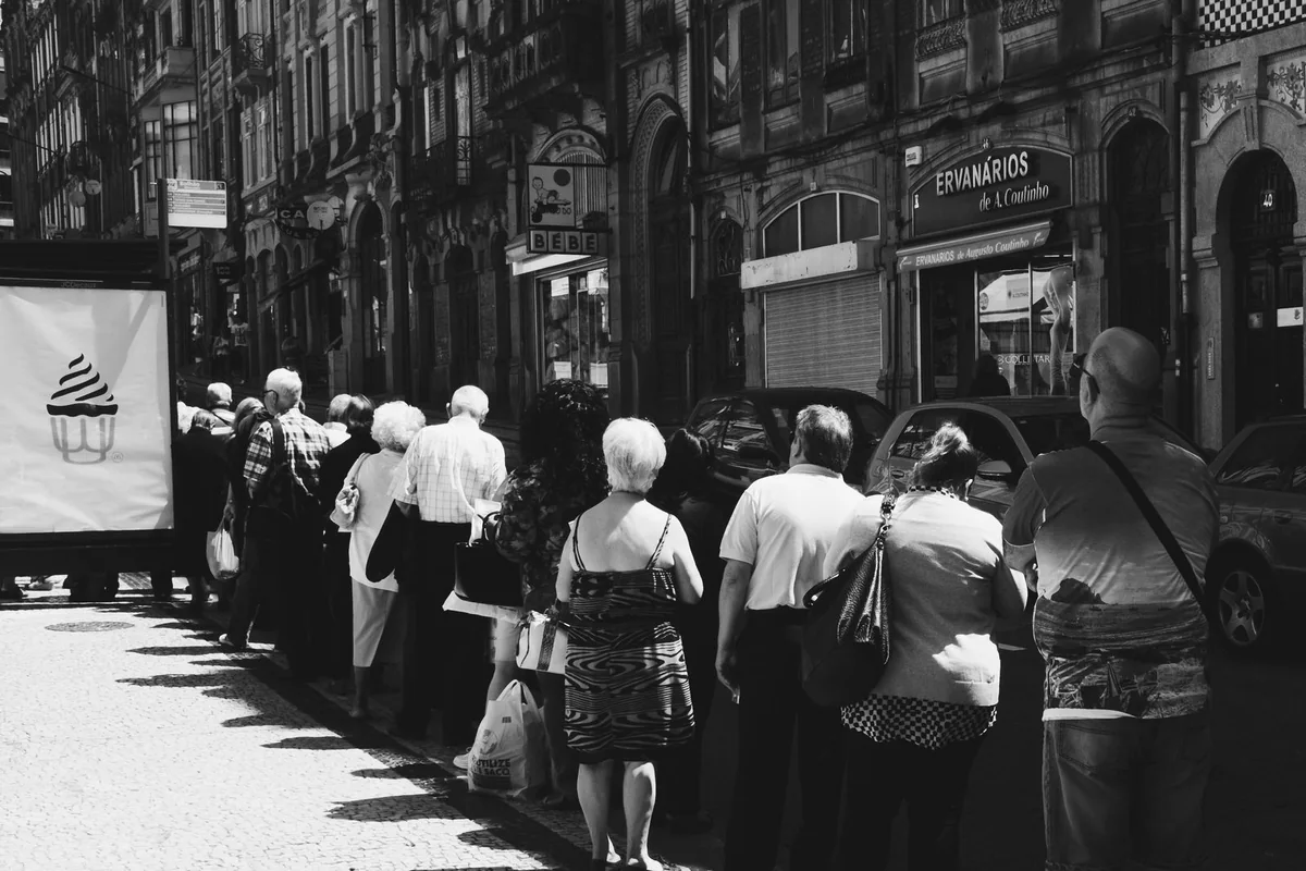 Black-and-white photograph of people waiting in a line along a city pavement.