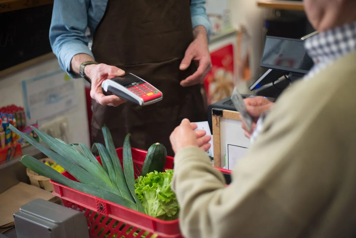 A customer pays by card beside a basket of groceries at a staffed checkout.