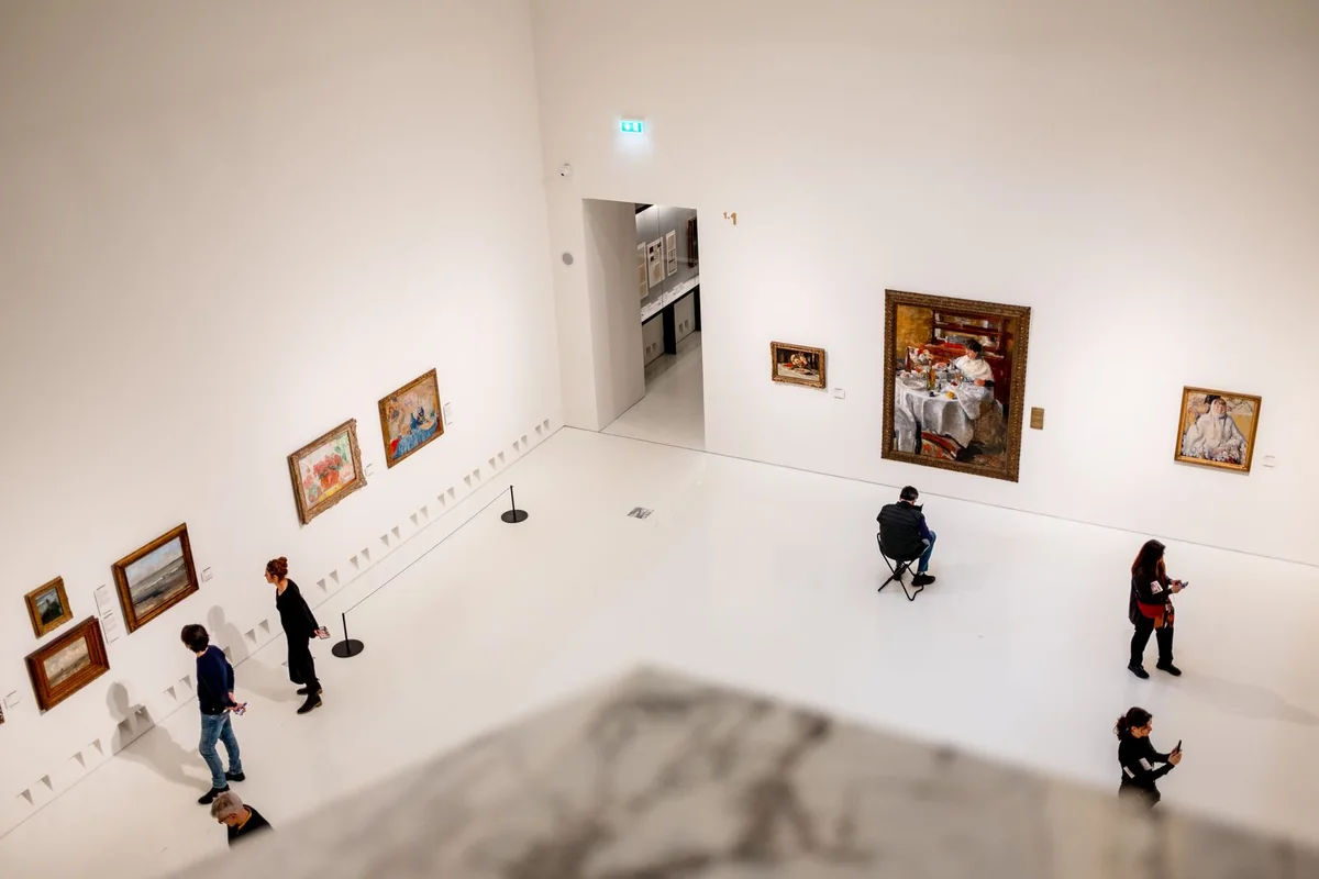 Visitors spread through a white art-gallery room, viewed from an upper level.