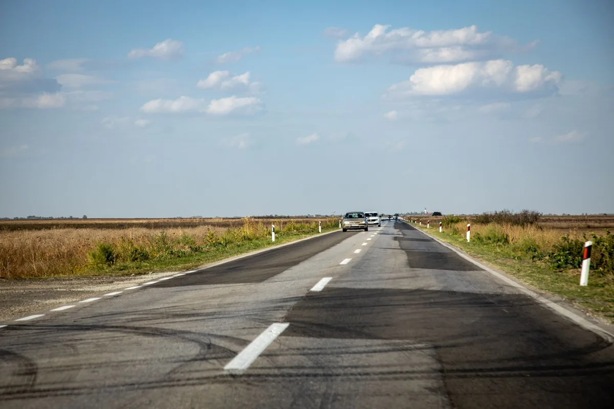 Straight rural road with vehicles in the distance, roadside markers and open fields.