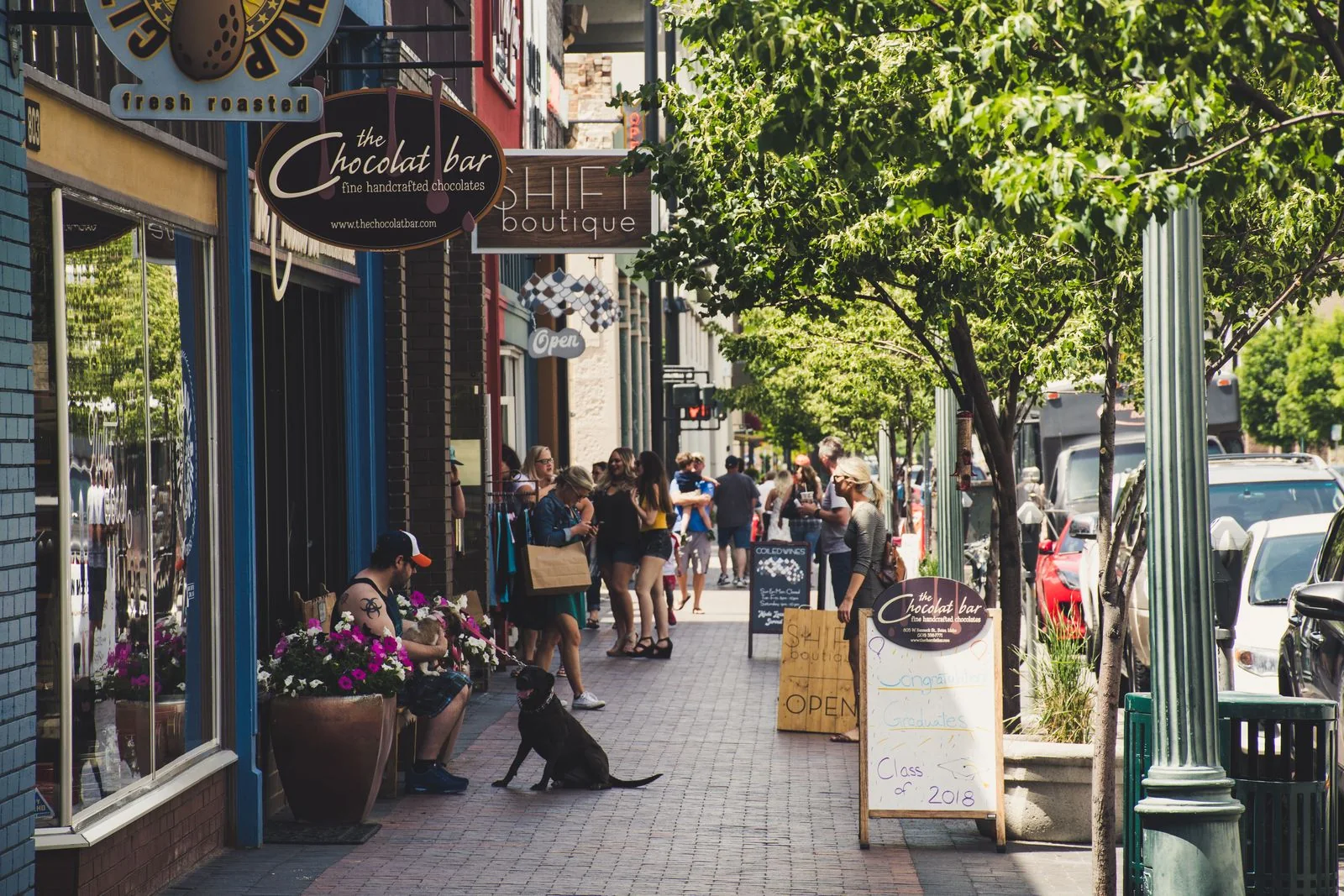 People walking and sitting along a tree-lined pavement outside independent shops.