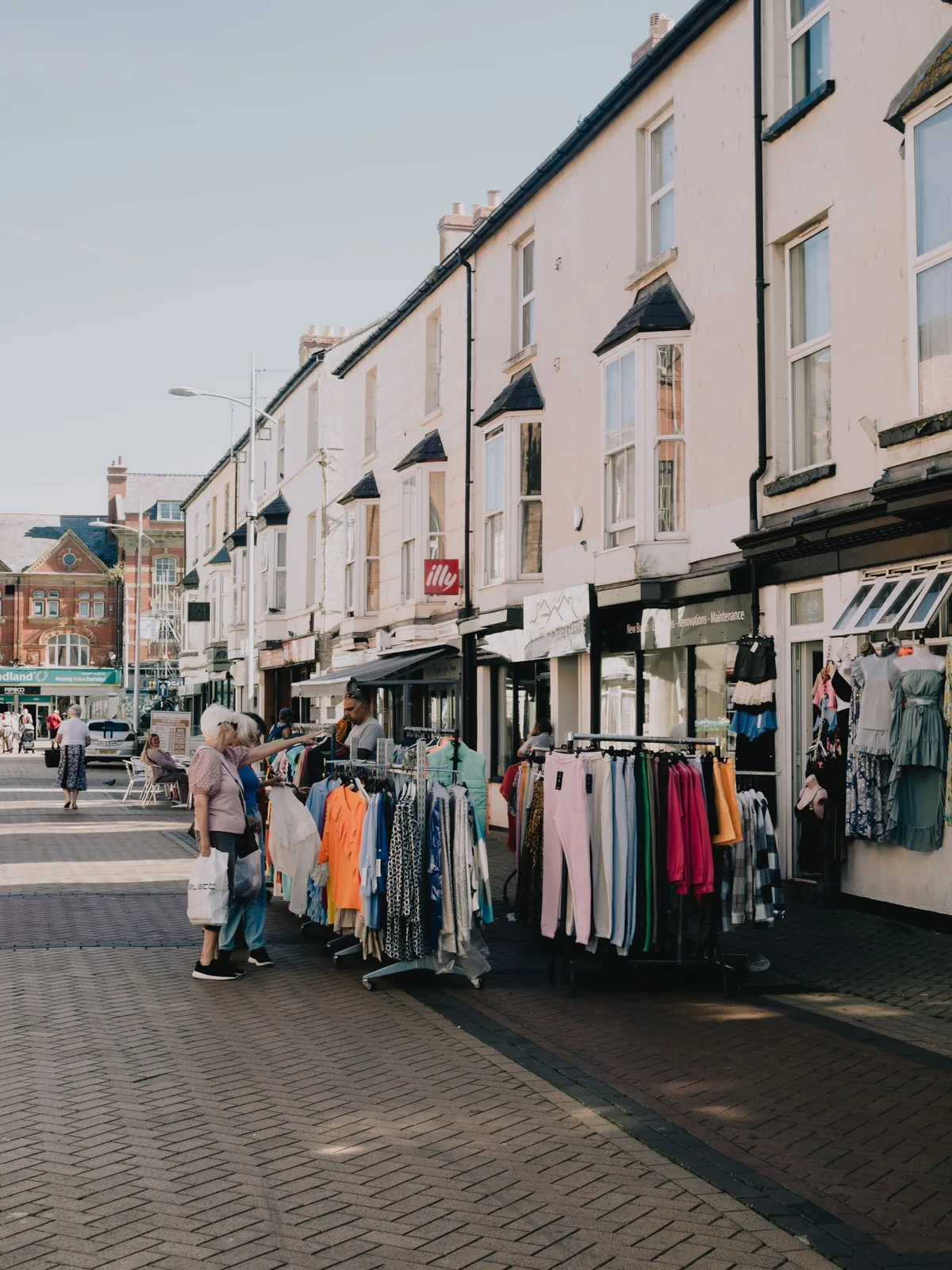A row of high-street shops with outdoor clothing racks and people browsing.
