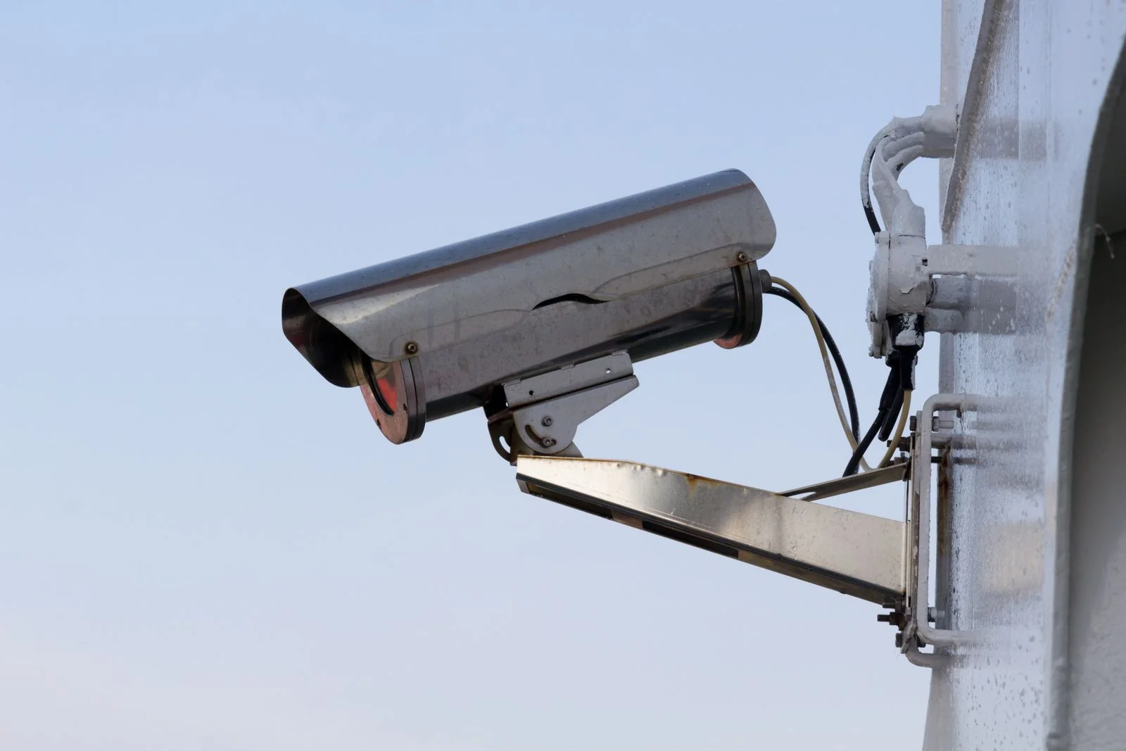 Outdoor surveillance camera and its metal mounting bracket against a clear sky.