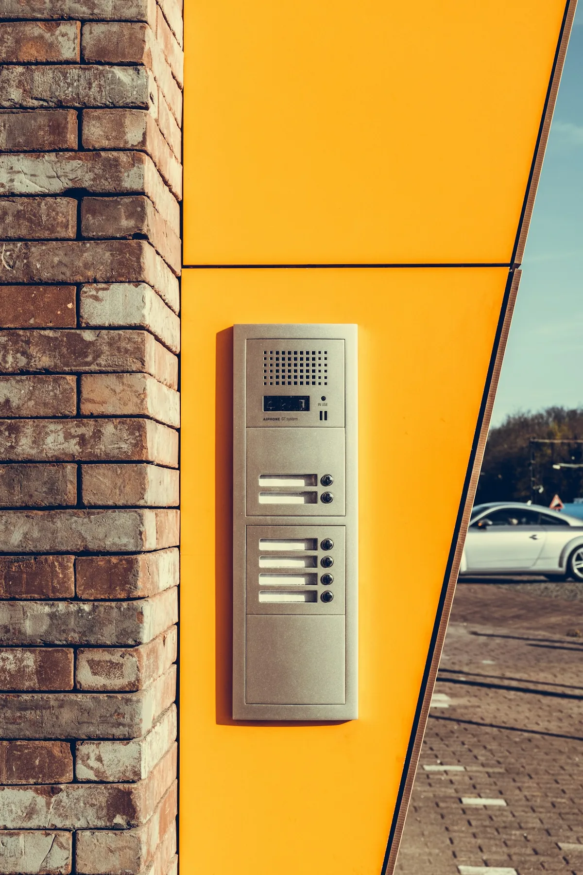 Metal entry intercom mounted beside brickwork on a yellow facade.