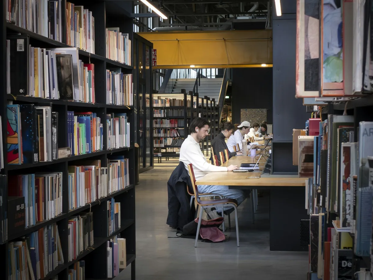 People studying at wooden tables between library bookshelves.