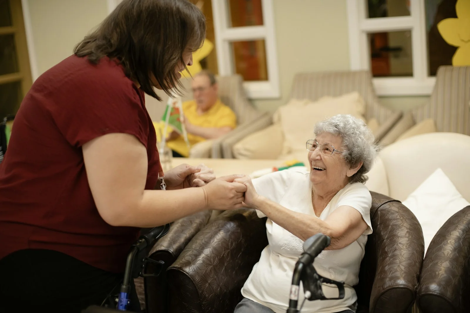 A caregiver assisting an older woman beside a walking frame in a shared room.