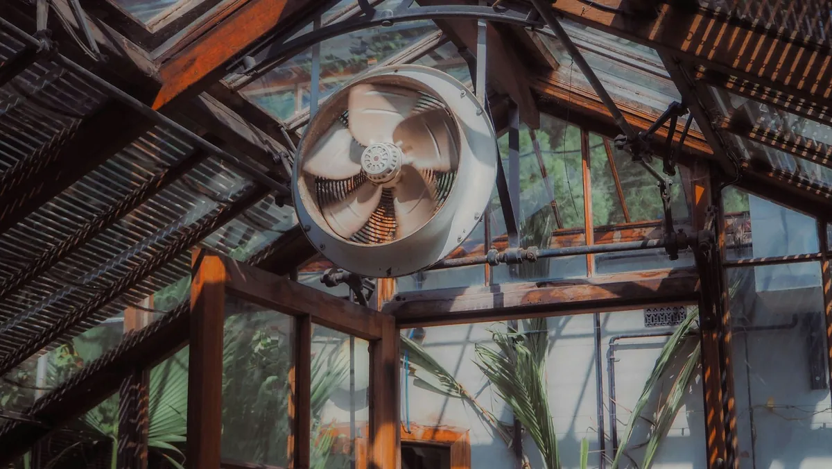 A ventilation fan mounted high inside a greenhouse roof structure.