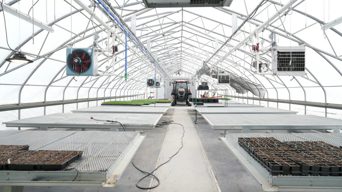 A long greenhouse aisle with growing tables, seedling trays, fans and suspended equipment.