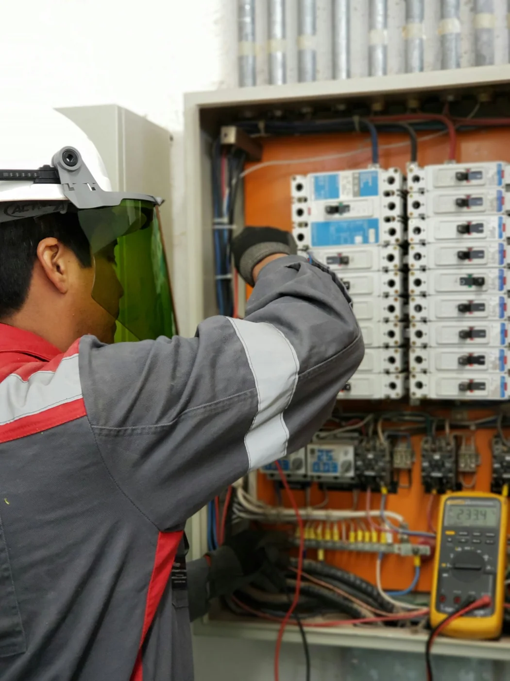 Technician wearing a hard hat and face shield working at an open electrical control cabinet.