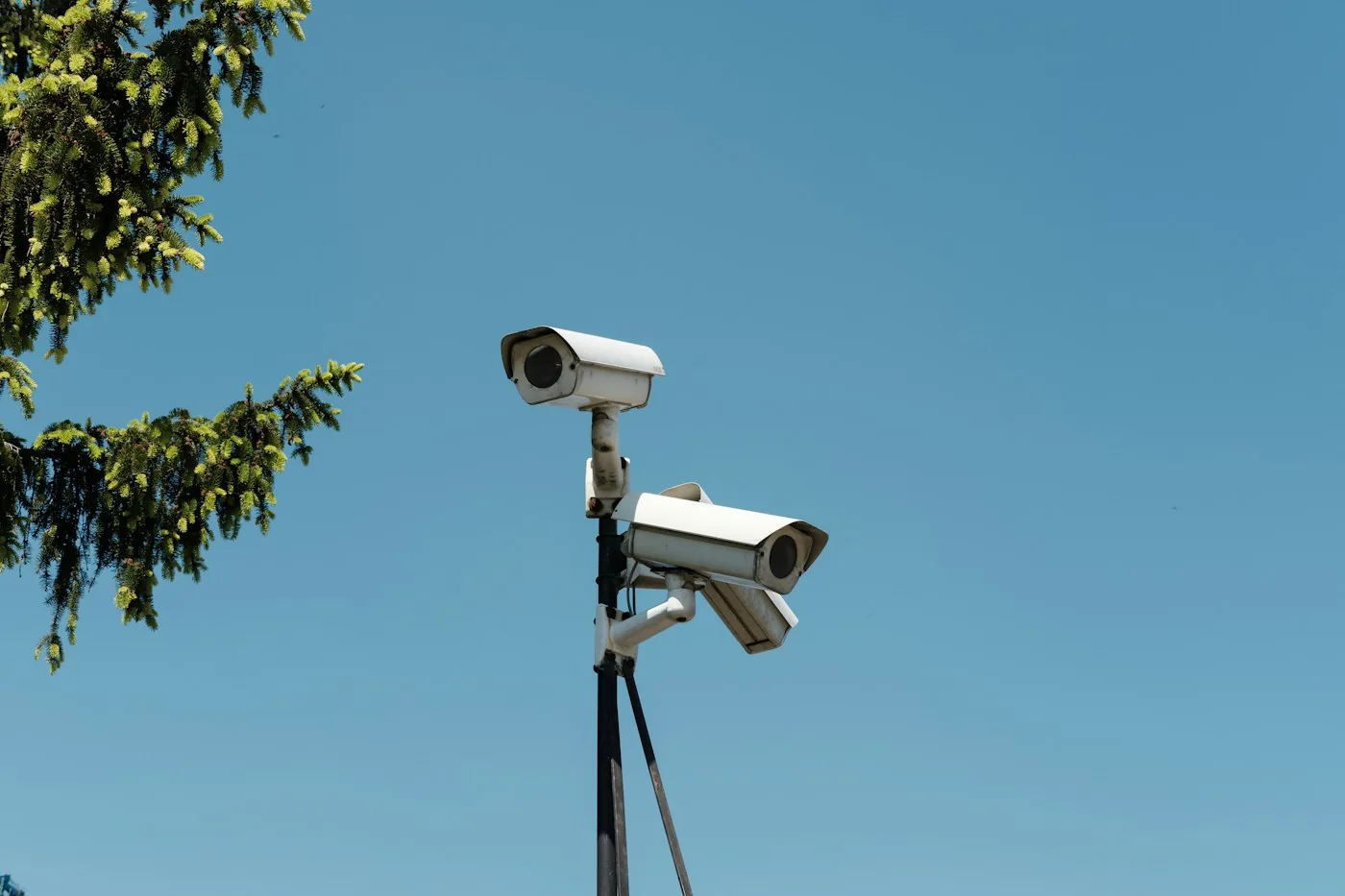 Two exterior surveillance cameras mounted on a pole against a blue sky.