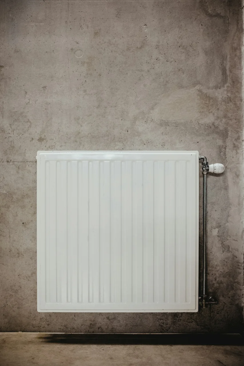 White panel radiator with a thermostatic valve and exposed pipework against a concrete wall.