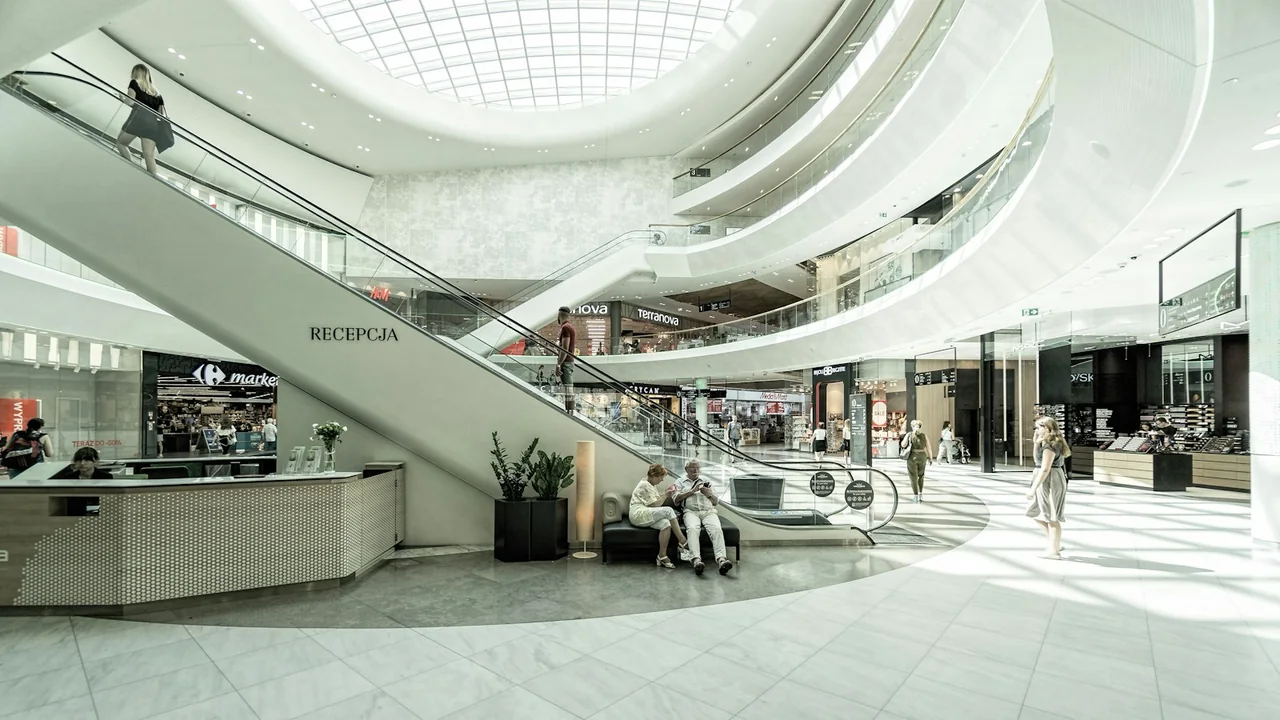 A shopping-centre atrium with stores, escalators and a glazed roof.