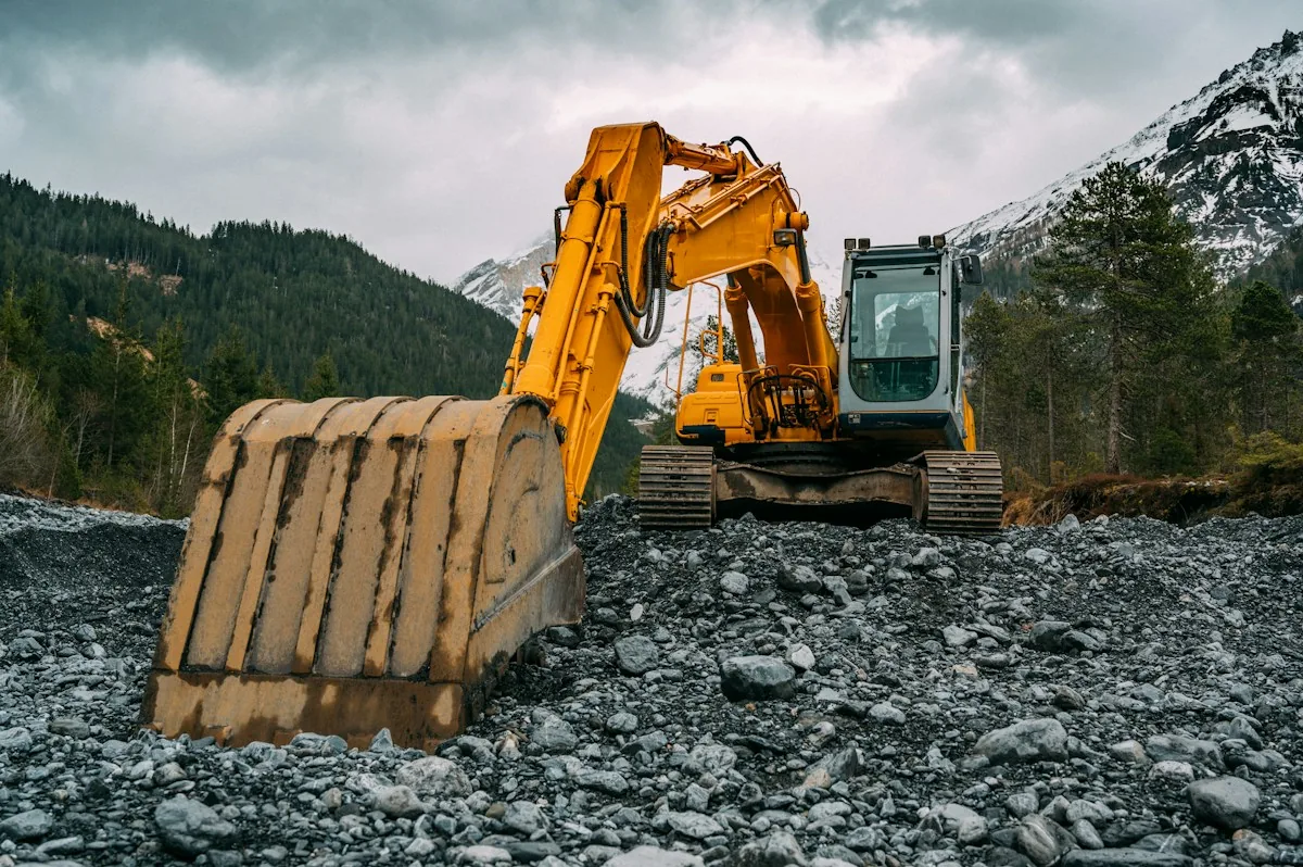 A yellow excavator on rocky ground with wooded mountains behind it.