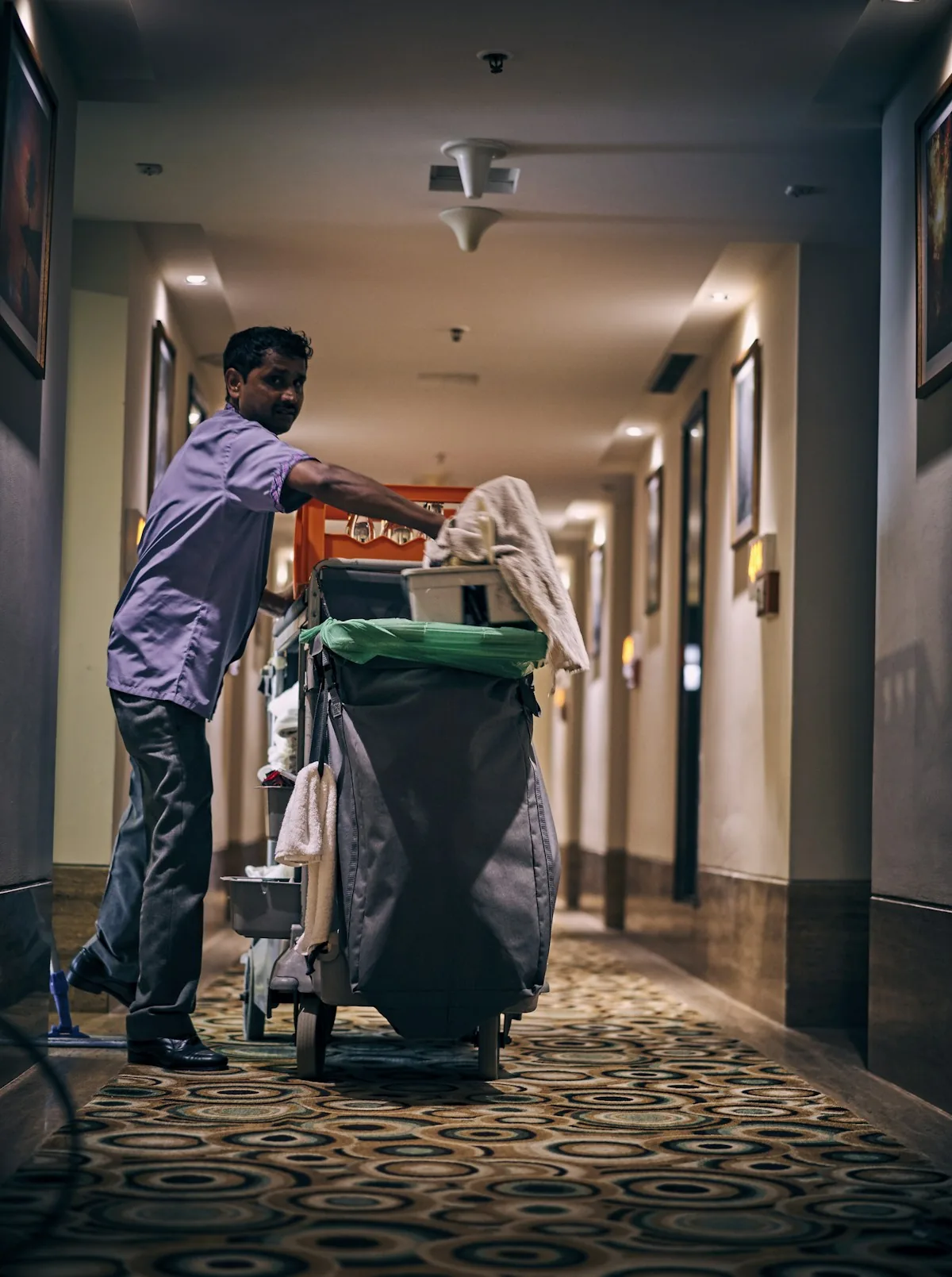 A cleaner moving a housekeeping cart through a hotel corridor.