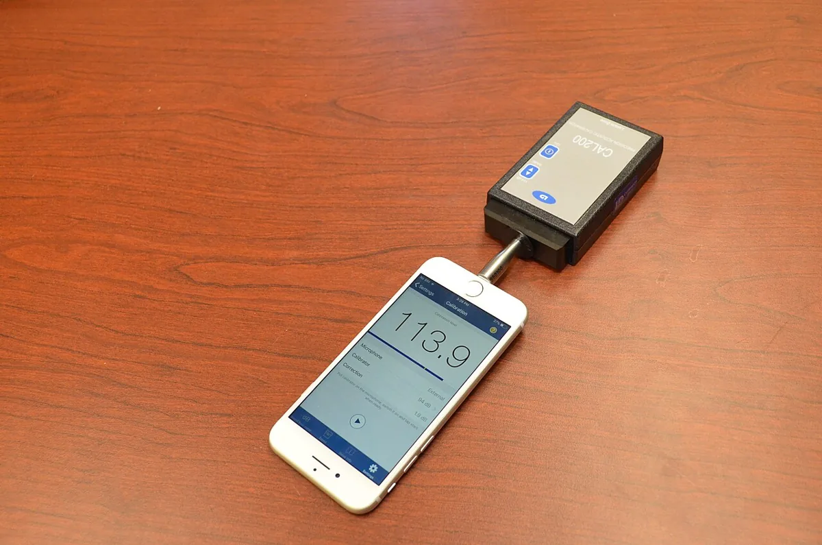A phone-based sound-level measurement setup connected to an acoustic calibrator on a wooden table.