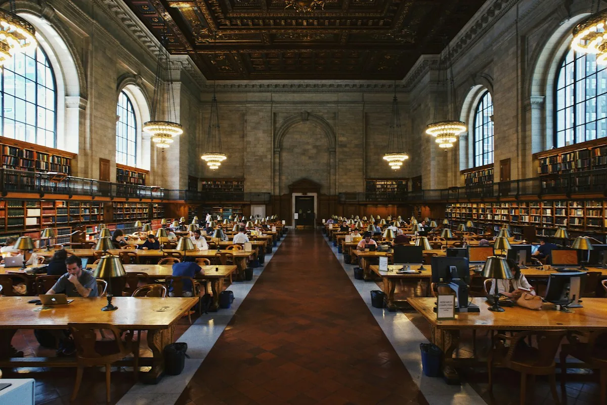 People study at parallel rows of desks in a large library reading room.
