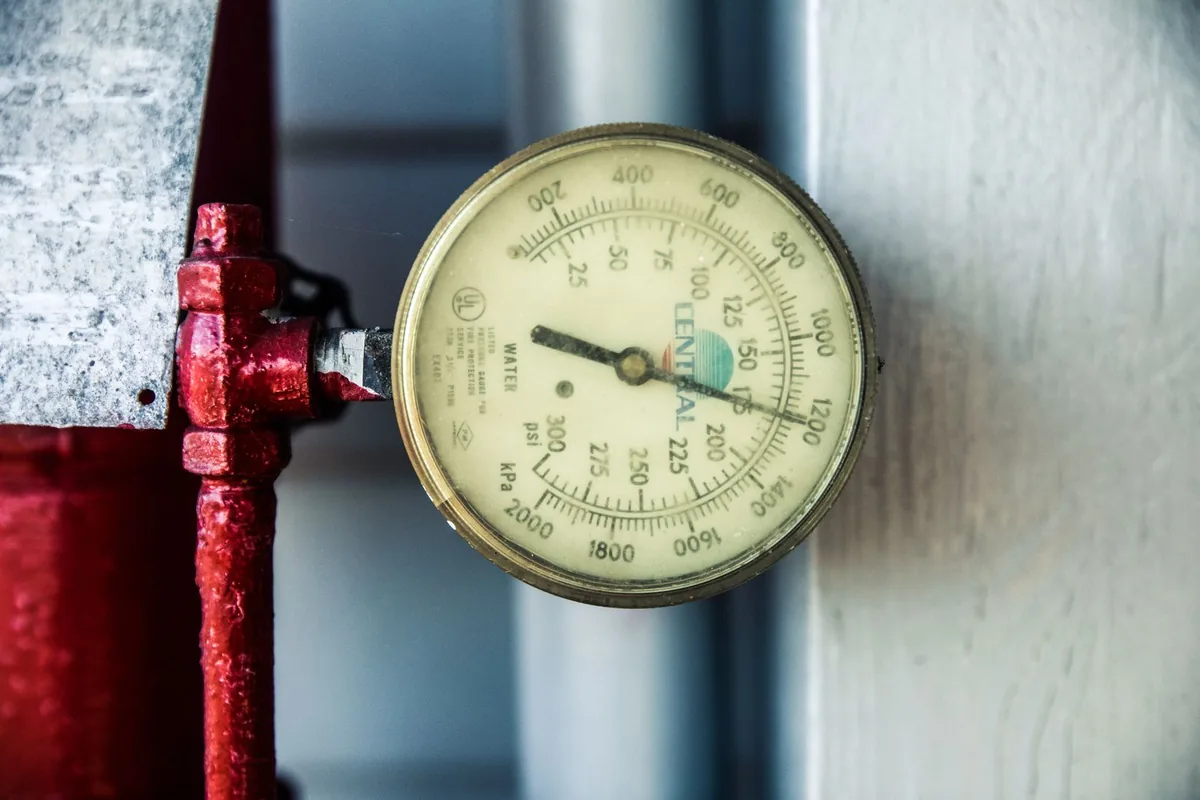 Round analog pressure gauge on red pipework beside a silver cylindrical surface.