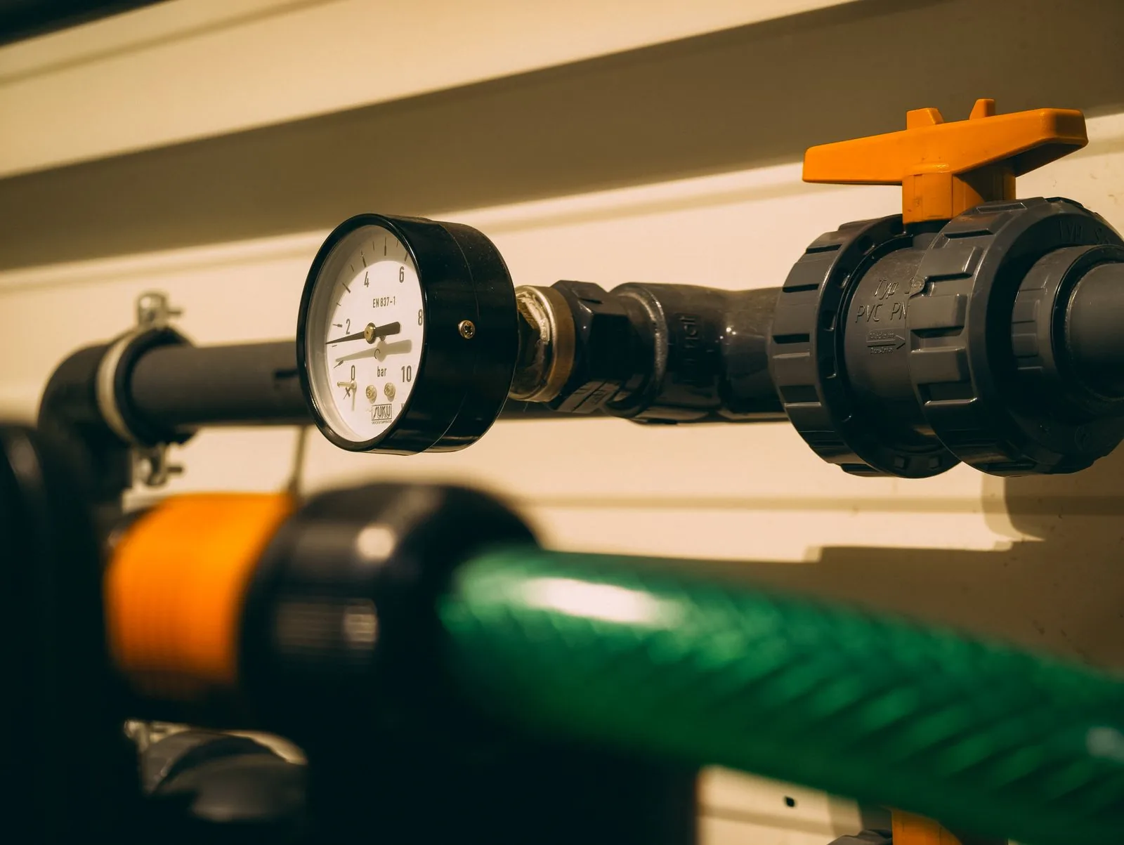 Analog pressure gauge fitted to dark pipework beside an orange isolation-valve handle.