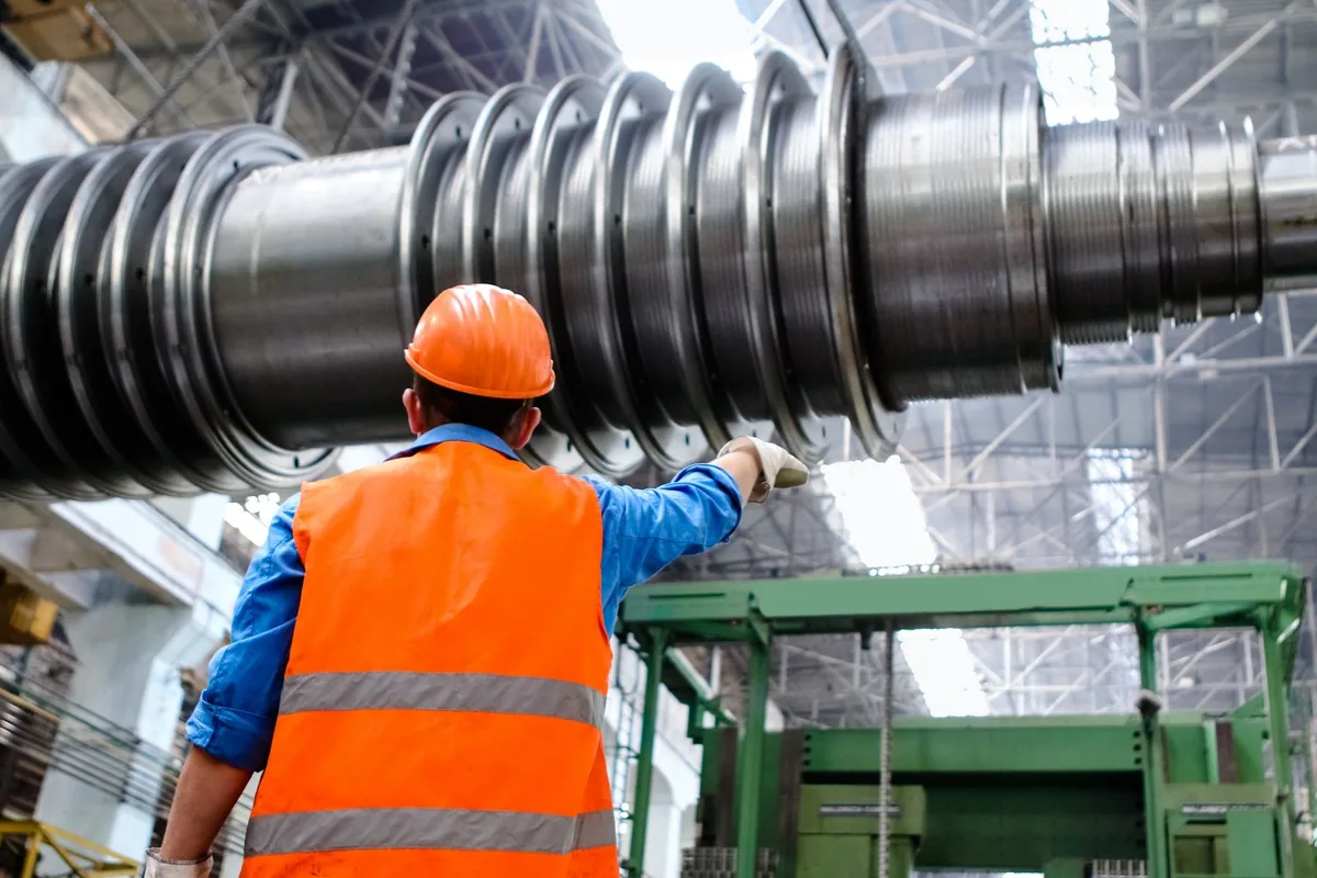 Worker wearing an orange hard hat and reflective vest beside a large metal component in a factory.