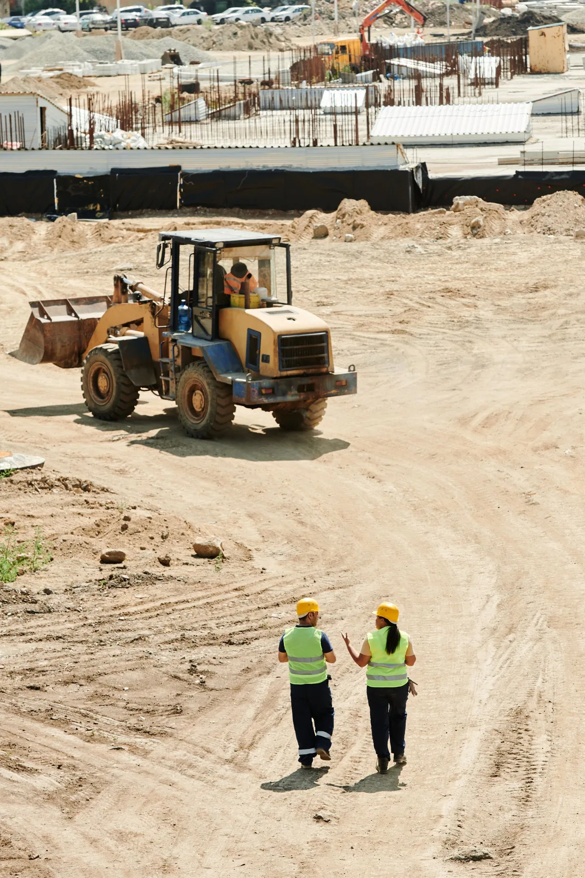 Two workers in high-visibility vests and hard hats walking across an open construction area near a backhoe.