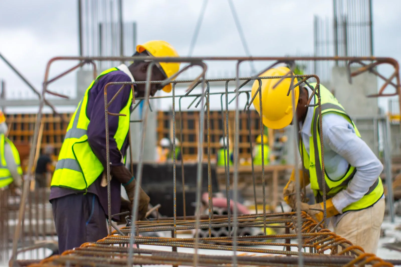 Two workers in yellow hard hats and high-visibility vests beside reinforcing steel at a construction site.