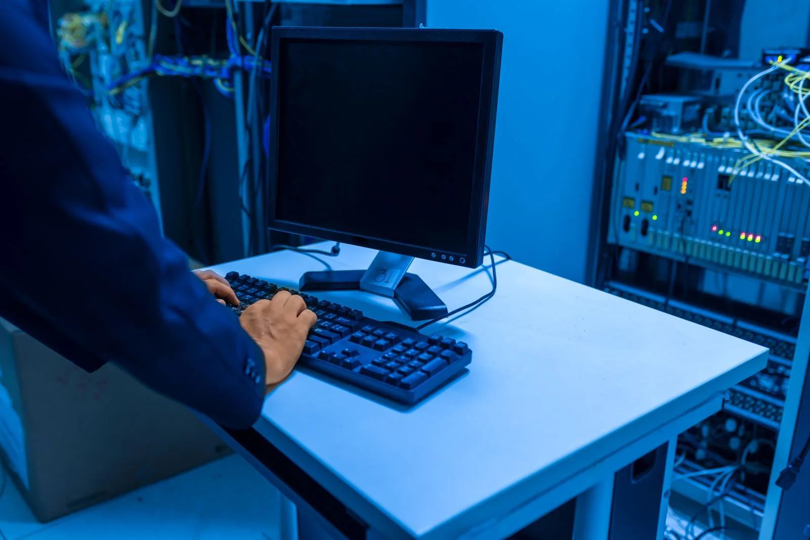 An IT technician uses a keyboard at a workstation beside network equipment cabinets.