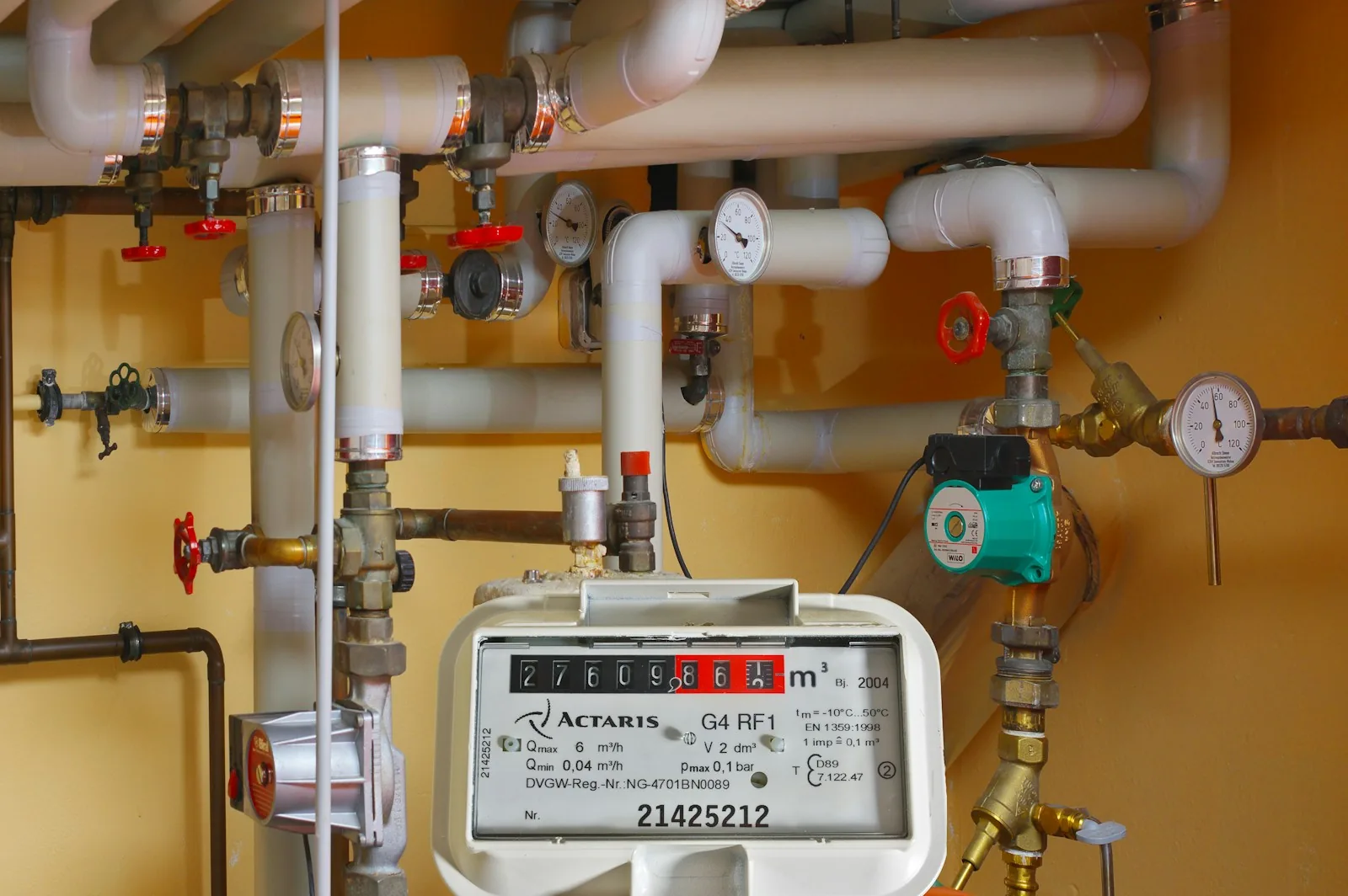 A mechanical utility meter in front of insulated pipework, valves and gauges in a plant room.
