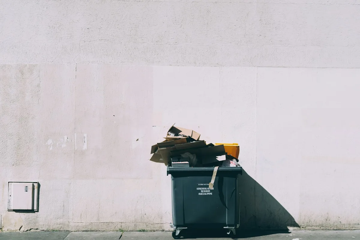 A wheeled waste container with cardboard protruding above its lid beside a pale wall.