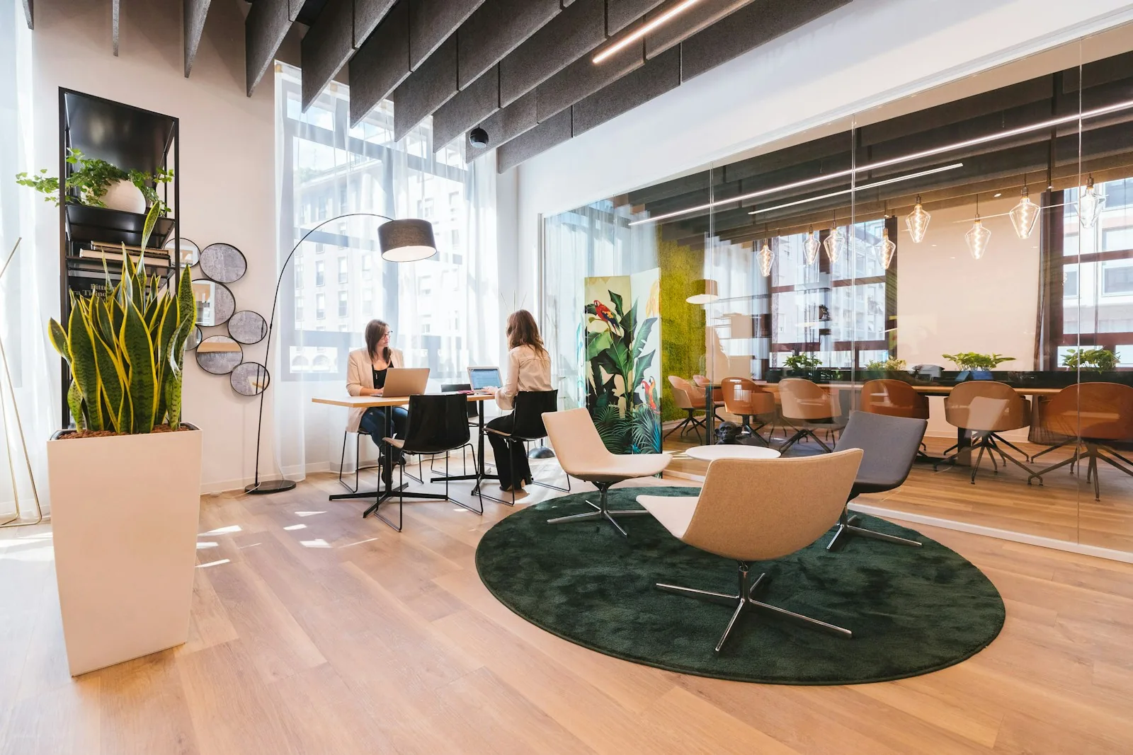 Two people work at a table beside lounge seating in a bright shared office.