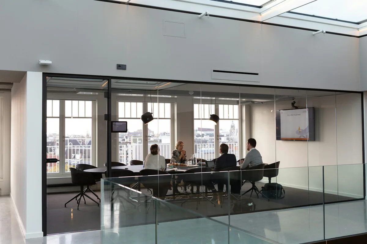 People meet around a table inside a glass-walled office meeting room.