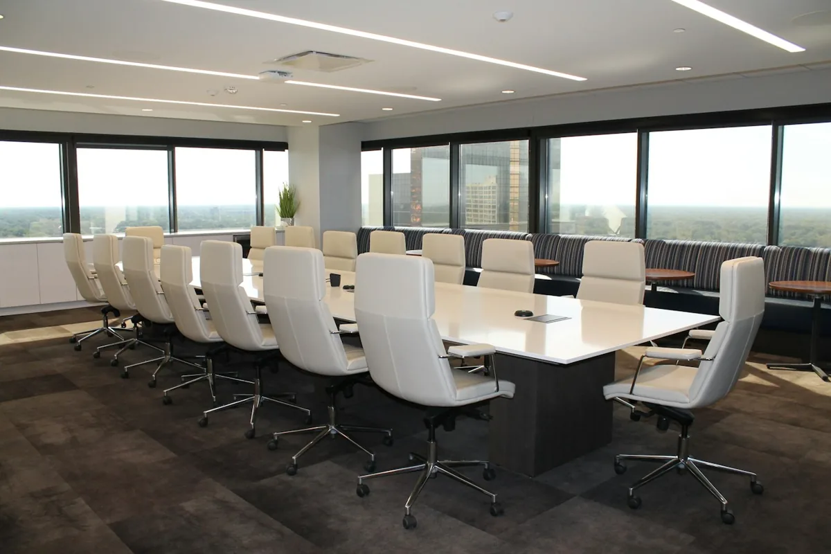 An empty meeting room with a long white conference table, white chairs and ceiling strip lights.