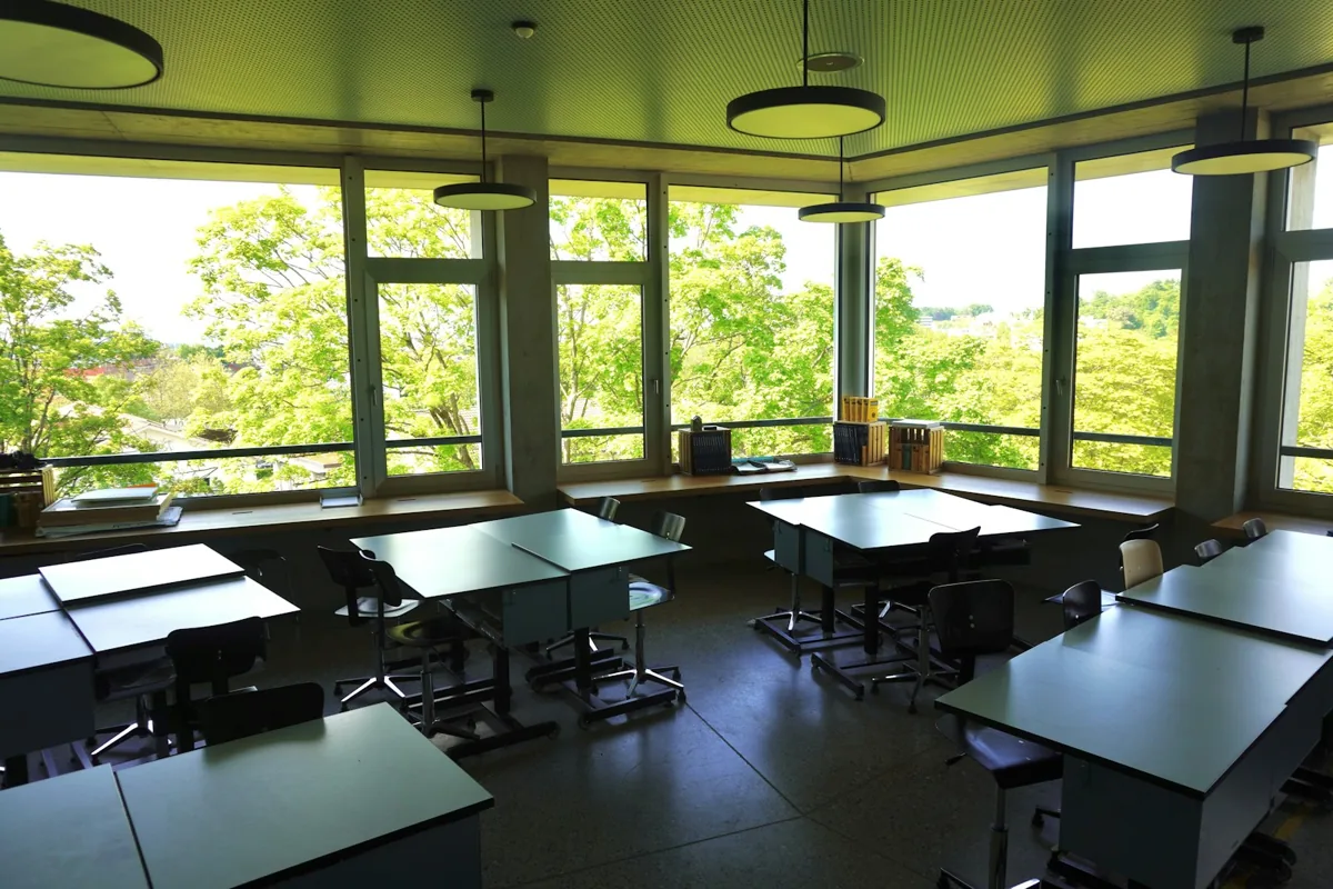 An empty classroom with desks, large windows and circular ceiling lights.