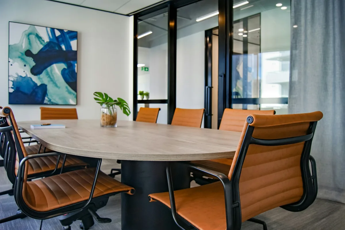 An empty glass-walled meeting room with a wooden table and brown chairs.
