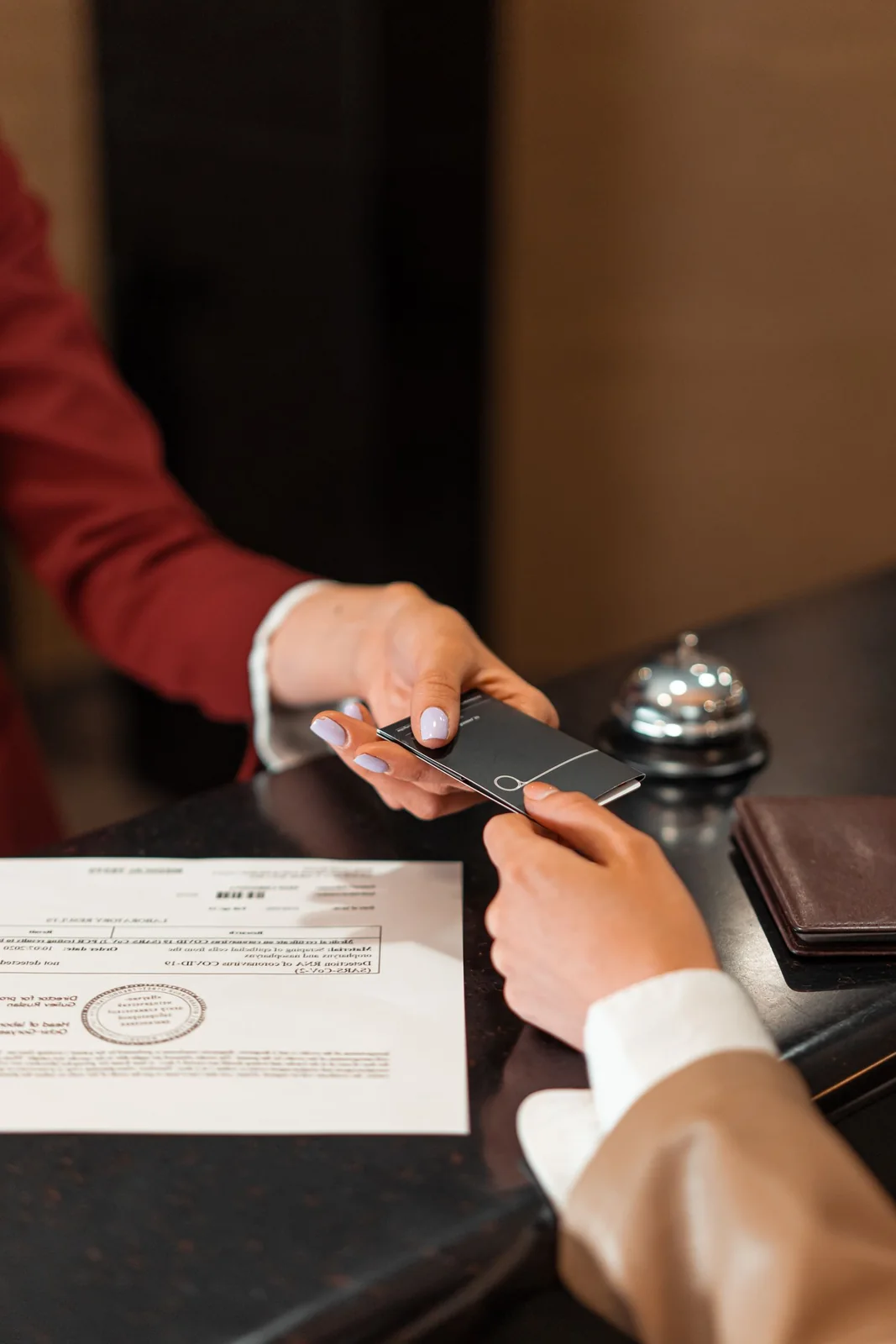 Hands exchange a hotel key card across a reception counter beside a bell and paperwork.