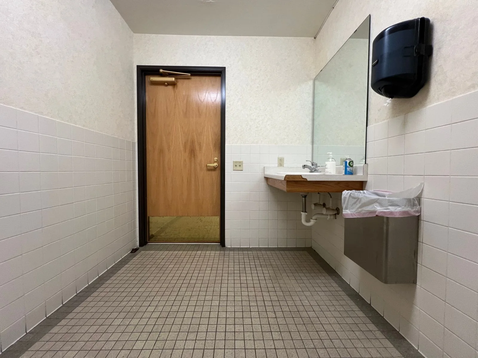 A wall-mounted paper dispenser above a washbasin and waste bin in a tiled restroom.