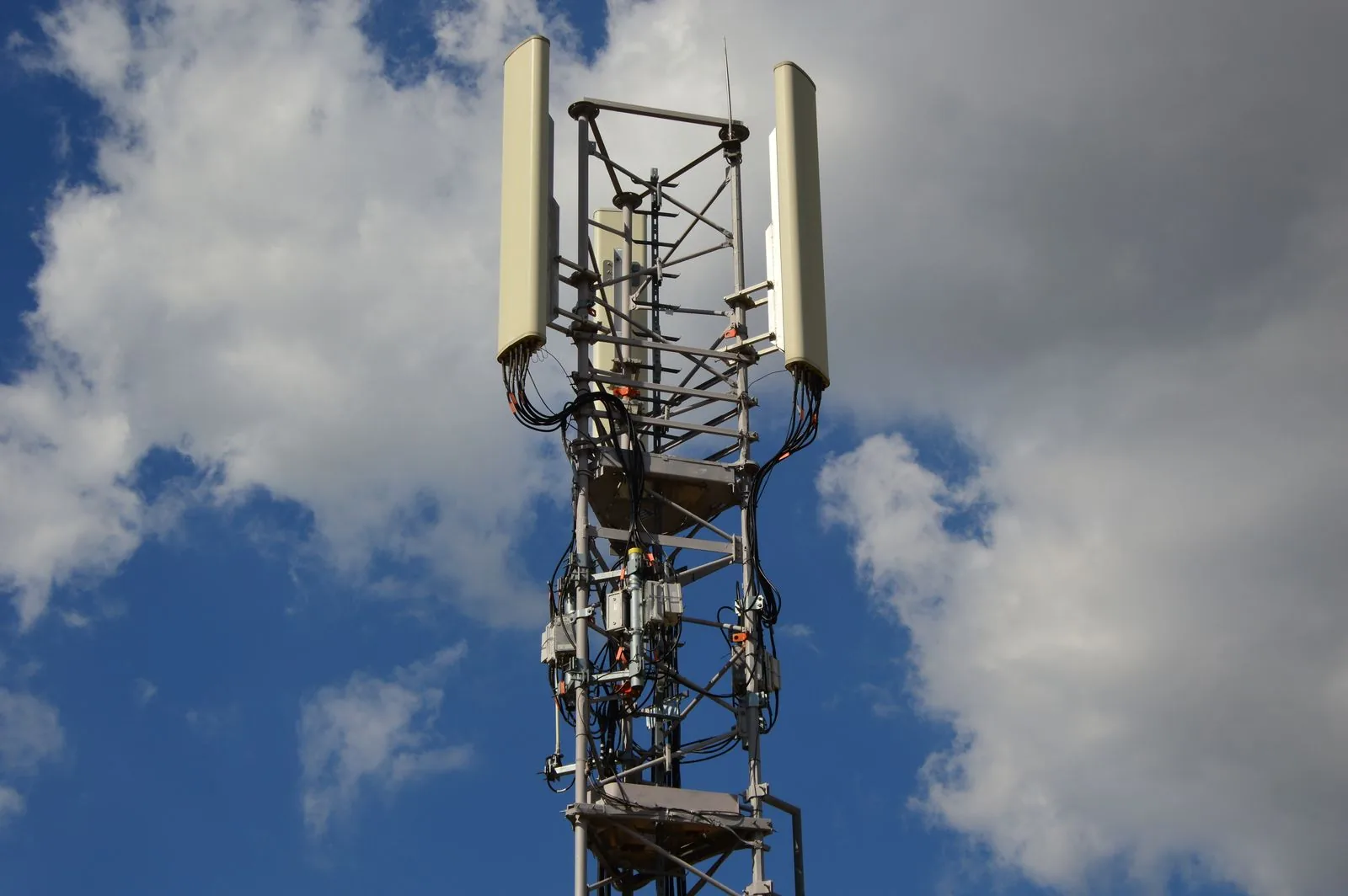 Cellular antennas and cabling on a steel tower beneath clouds.