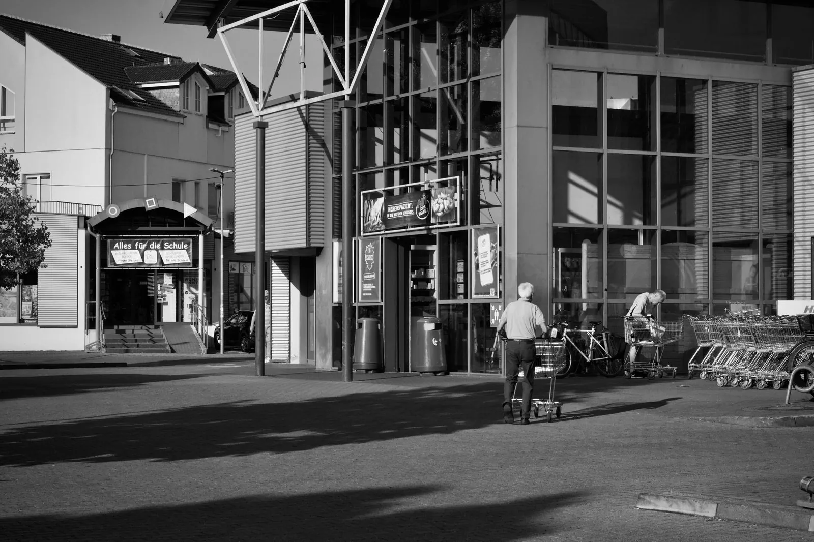 Shoppers and shopping carts outside a glass-fronted retail entrance in a black-and-white photograph.