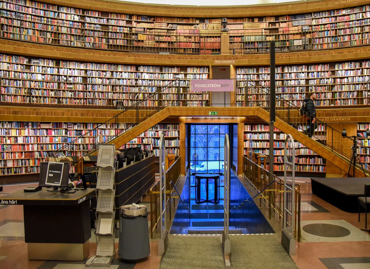 Library entrance beneath a stairway, surrounded by filled bookshelves, a circulation desk and visitors.