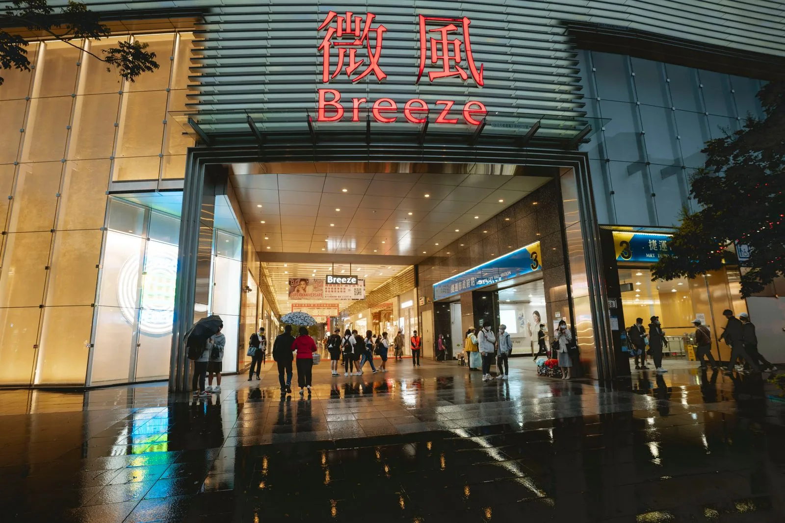 People walking and standing at a broad shopping-mall entrance on a rainy evening.